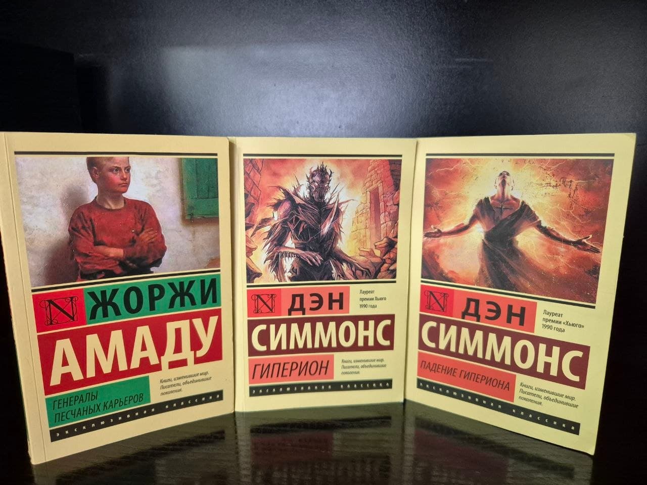 Три книги: "Генералы песчаных карьеров" Жоржи Амаду, "Гиперион" и "Падение Гипериона" Дэна Симмонса на тёмной поверхности.