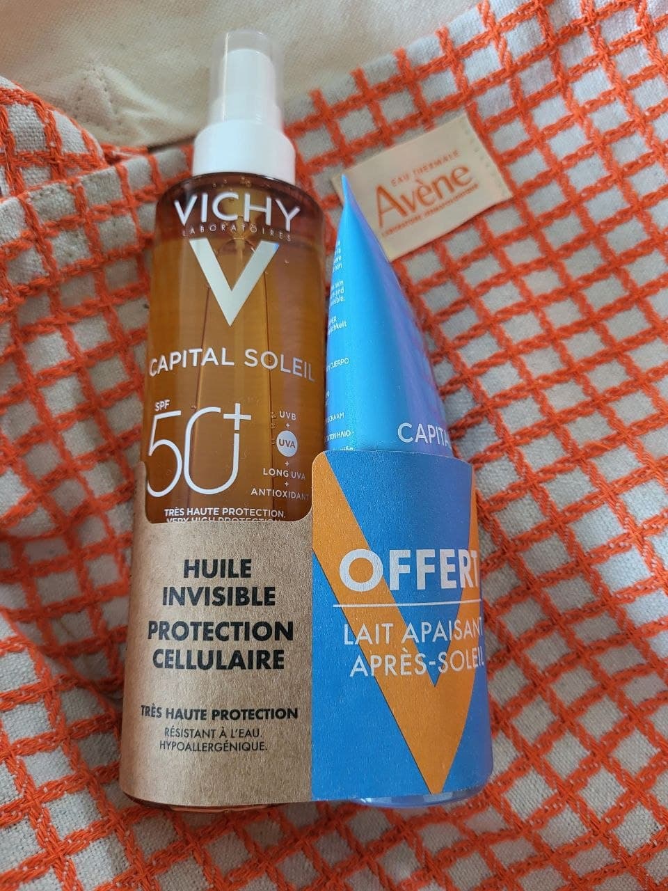 Солнцезащитный спрей Vichy Capital Soleil SPF 50+ и синий тюбик лосьона после загара на оранжевой ткани.