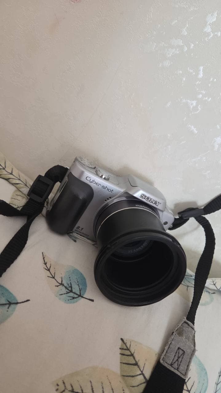 Цифровой фотоаппарат Sony Cyber-shot DSC-H3 с объективом и ремешком на фоне ткани с рисунком листьев.