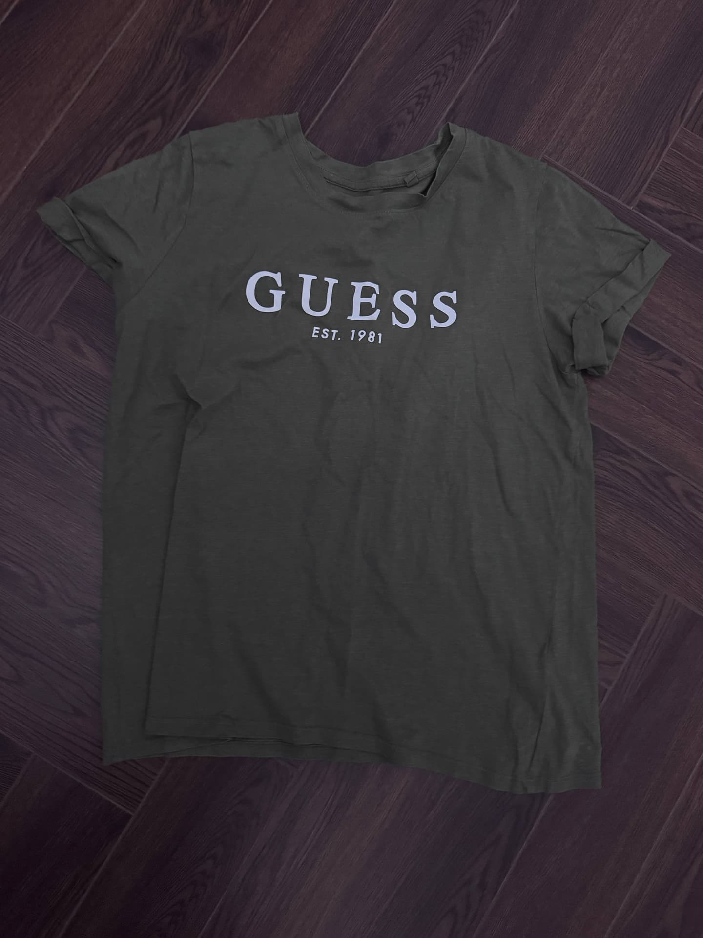 Темно-зеленая футболка с надписью GUESS EST. 1981 на темном деревянном полу.