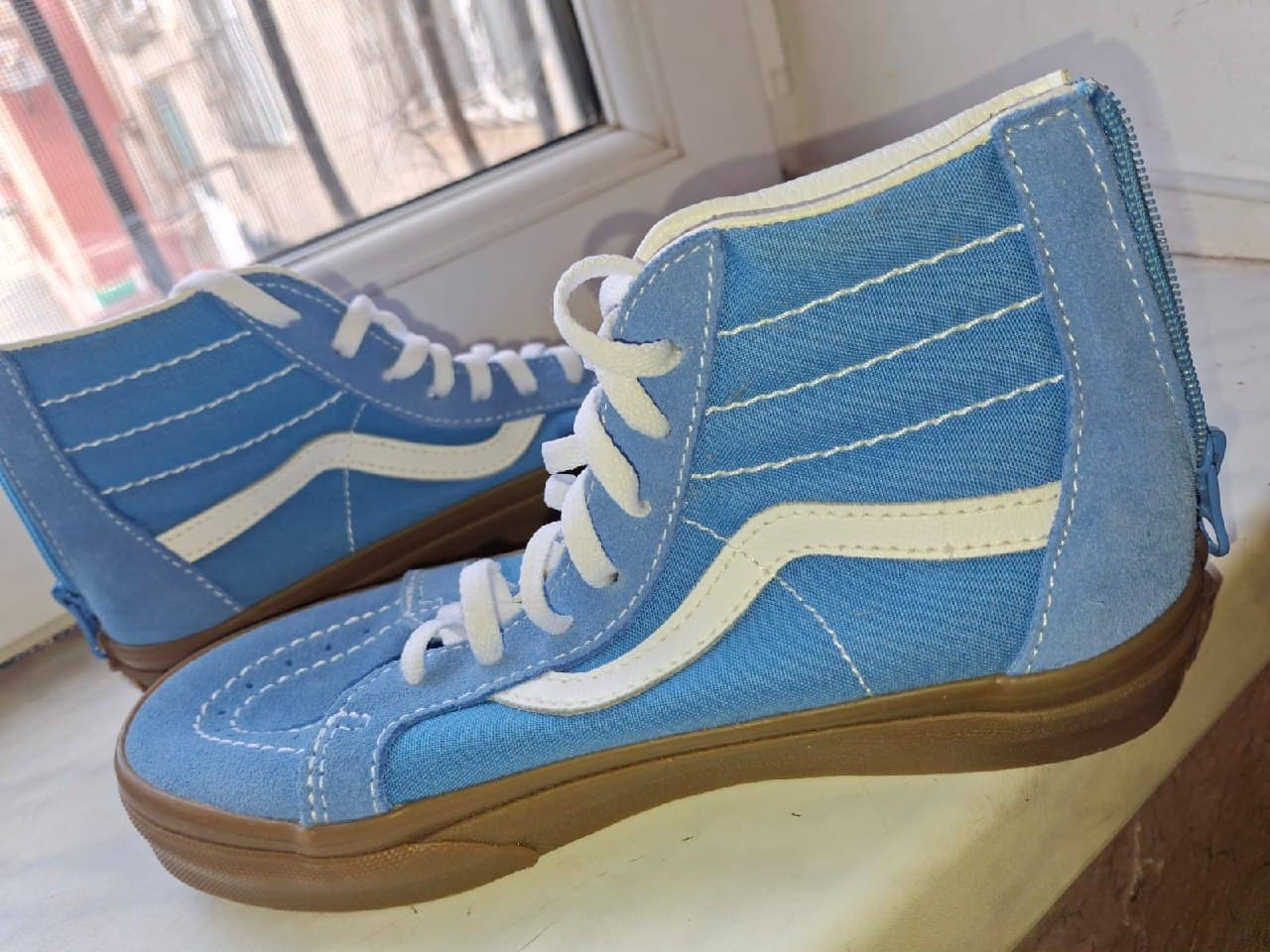 Голубые высокие кроссовки Vans с белыми шнурками и коричневой подошвой на белой поверхности у окна.