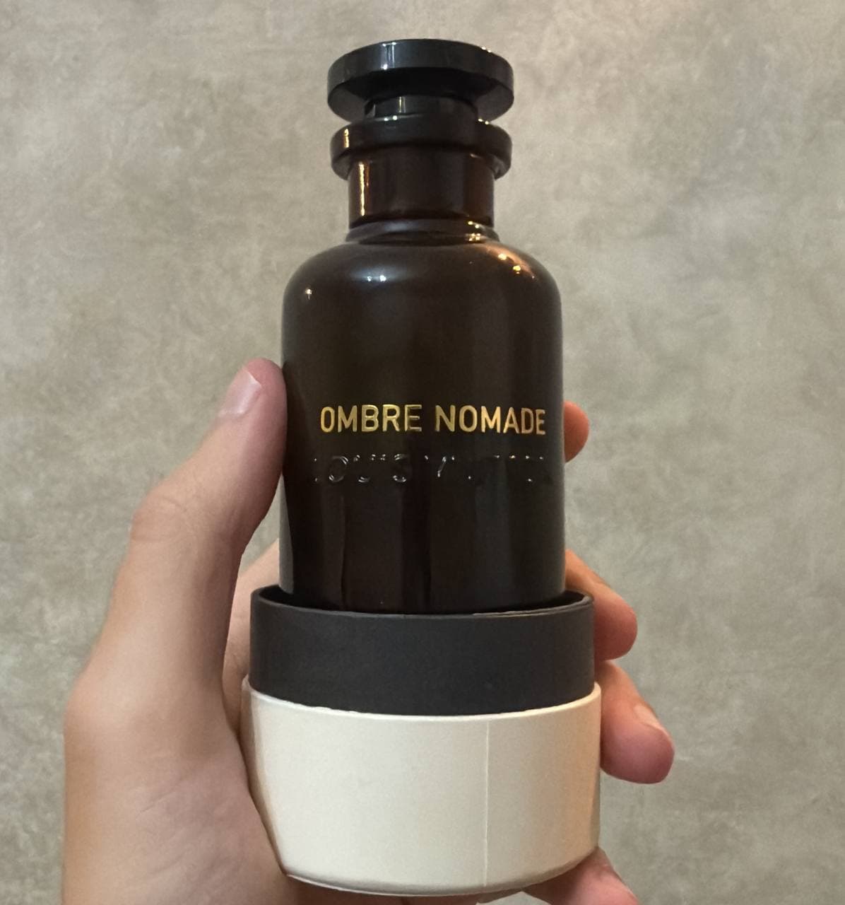 Темная бутылка духов с надписью OMBRE NOMADE в руке на светлом фоне.