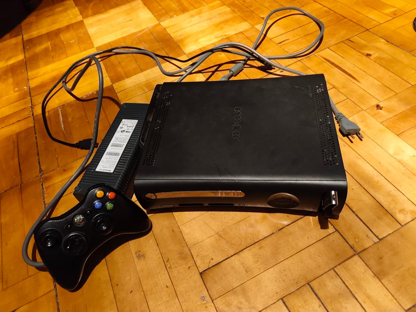 Игровая консоль Xbox 360 с одним контроллером и кабелями на деревянном полу.