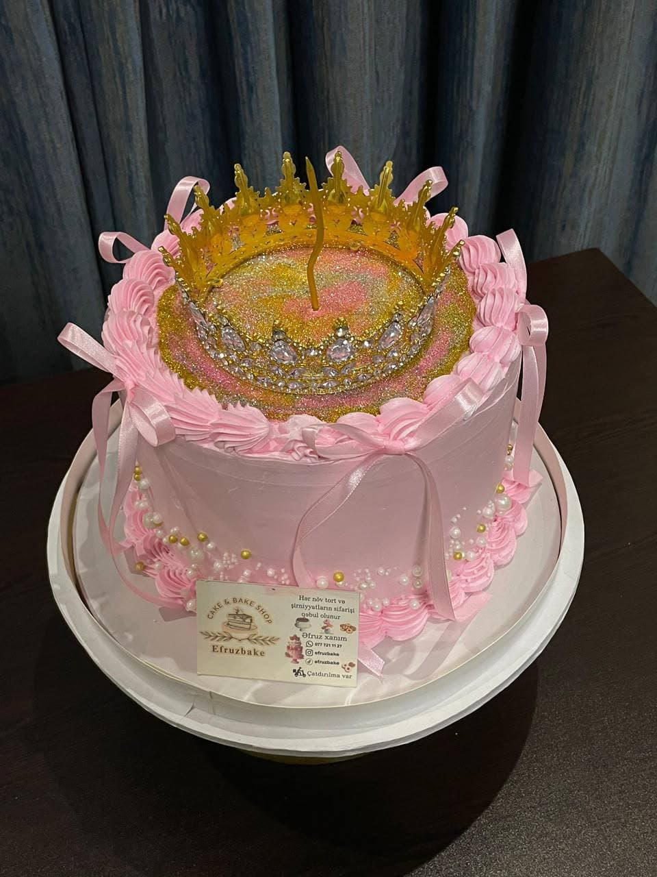 Розовый торт с золотой короной и свечой сверху, украшенный лентами и бусинами.