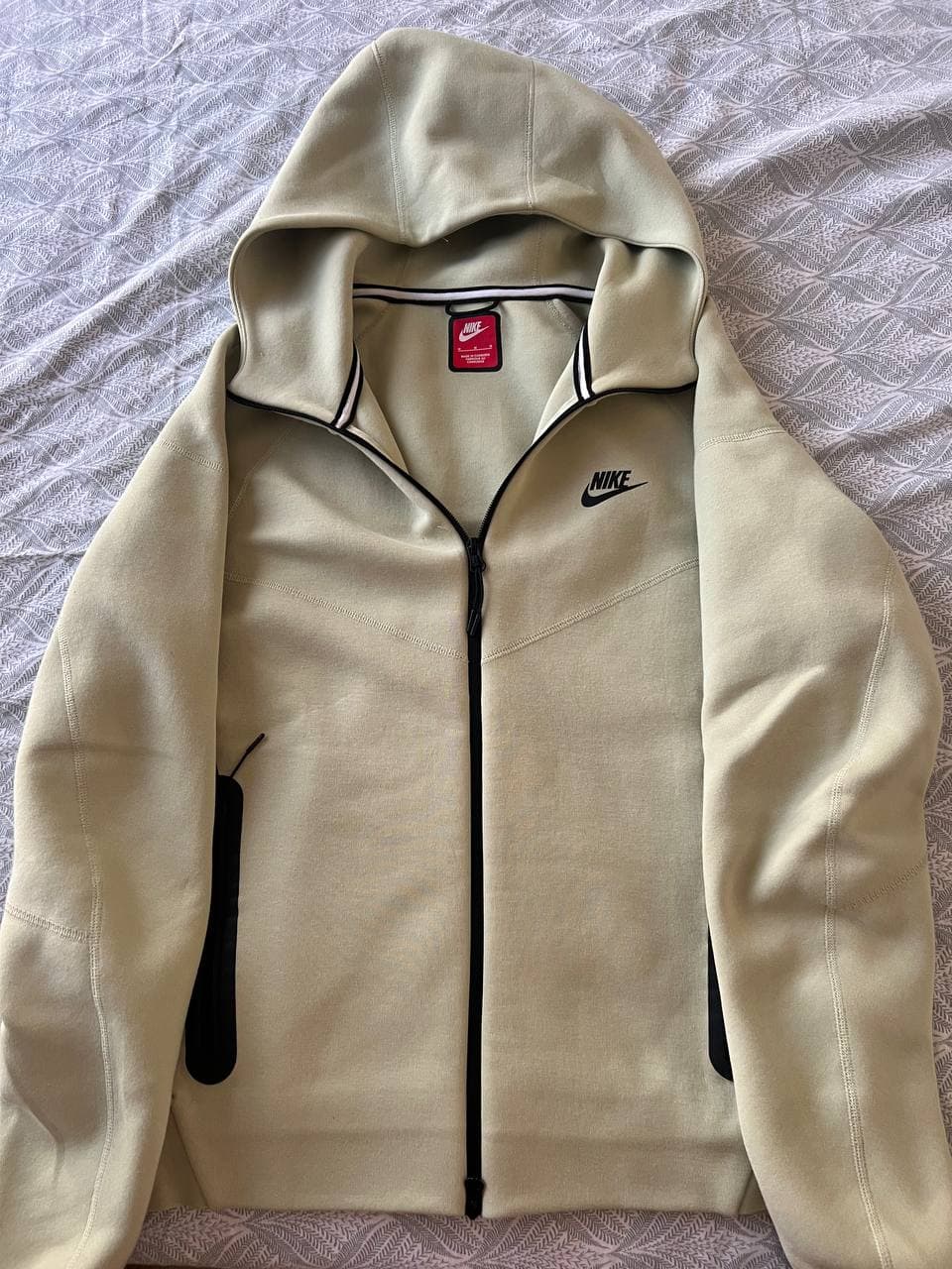 Светлая куртка-худи Nike с капюшоном и молнией, лежащая на покрывале.