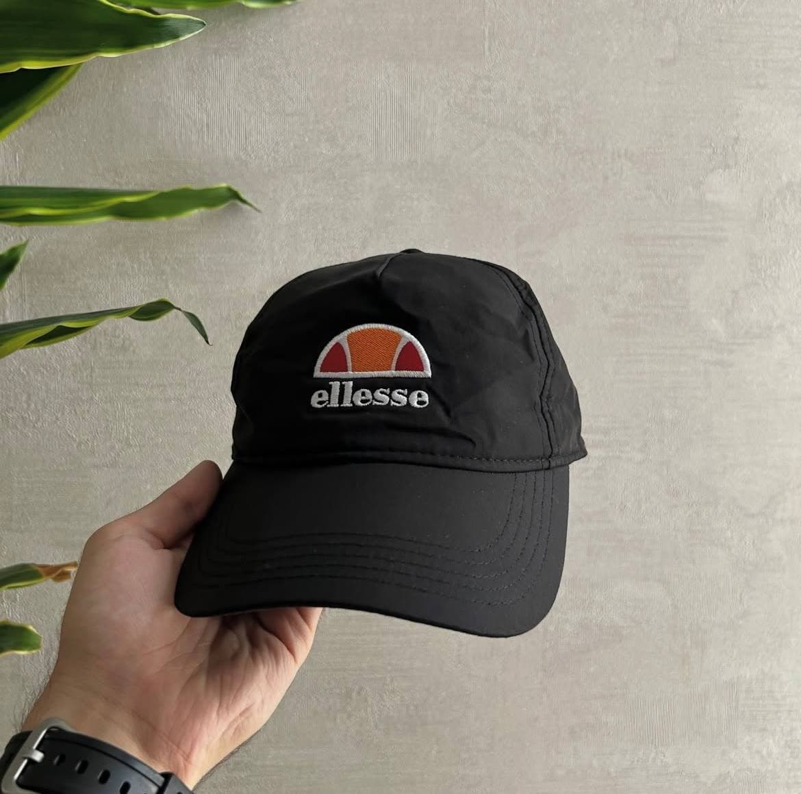 Черная бейсболка с логотипом ellesse, держится в руке на фоне стены и растения.