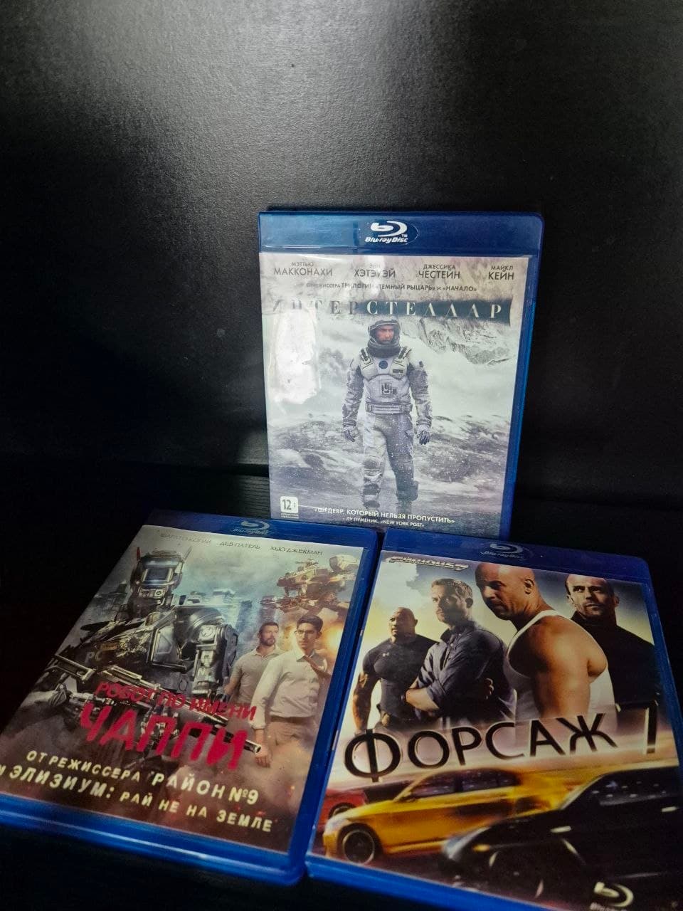 Три Blu-ray диска с фильмами "Интерстеллар", "Робот по имени Чаппи" и "Форсаж 7" на черном фоне.