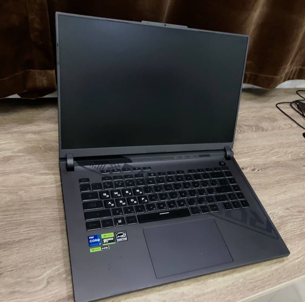Открытый ноутбук Asus ROG Strix G16 на деревянном столе с выключенным экраном и видимой клавиатурой.