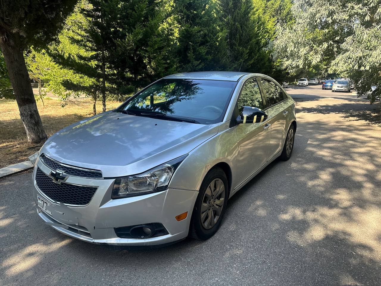Серебристый Chevrolet Cruze 2014 года, вид спереди слева на фоне деревьев и дороги.