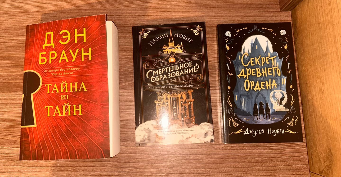 Три книги на деревянном столе: "Тайна из тайн" Дэн Браун, "Смертельное образование" Наоми Новик, "Секрет древнего ордена" Джулии Ноубел.