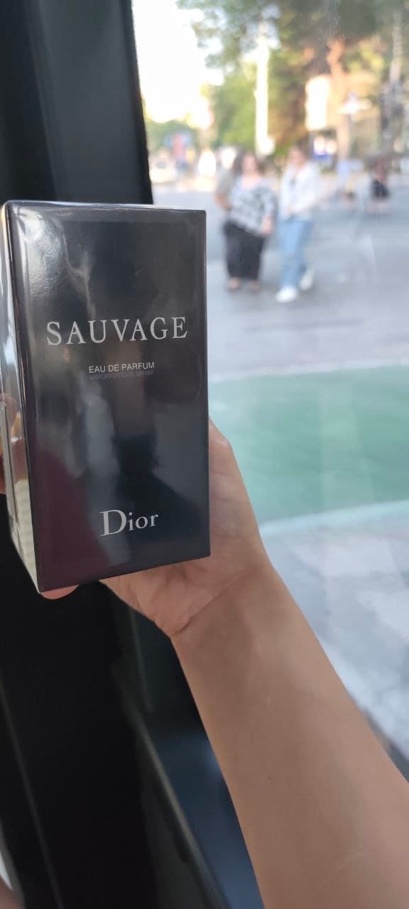 Коробка парфюма Dior Sauvage в руке на фоне улицы.